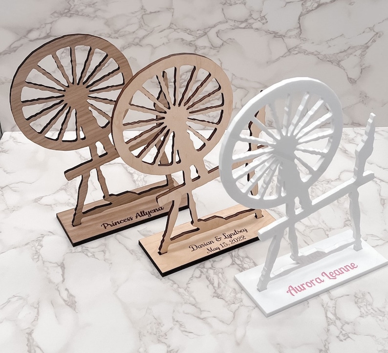 May include: Three wooden spinning wheels with personalized names. The first wheel is natural wood with the name "Princess Allyeana" engraved on the base. The second wheel is natural wood with the name "Dorian & Lyndsey May 15, 2022" engraved on the base. The third wheel is white with the name "Aurora Jeanne" engraved on the base.