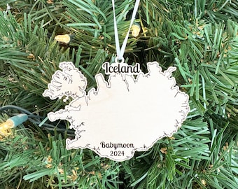 May include: Wooden ornament shaped like the outline of Iceland with the text "Iceland" and "Babymoon 2024" on it. The ornament is hanging on a Christmas tree.