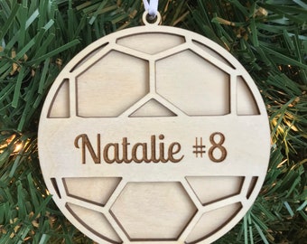 May include: A wooden soccer ball ornament with a cutout design. The ornament is personalized with the name "Natalie #8" in a brown font.