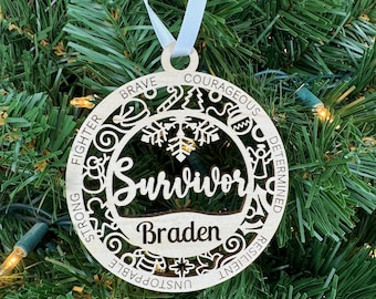 May include: A wooden Christmas ornament with the word "Survivor" and the name "Braden" inscribed. The ornament is decorated with a snowflake and the words "Brave, Fighter, Strong, Unstoppable, Resilient, Determined, Courageous".