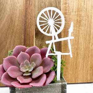 May include: White wooden spinning wheel cutout, a decorative accent for a succulent planter. The wheel is on a white stick, and the planter is filled with a pink succulent.