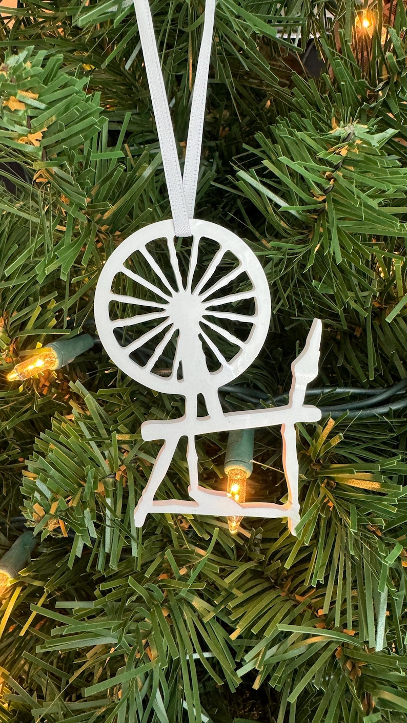 May include: White spinning wheel Christmas ornament hanging on a green artificial Christmas tree with white lights.
