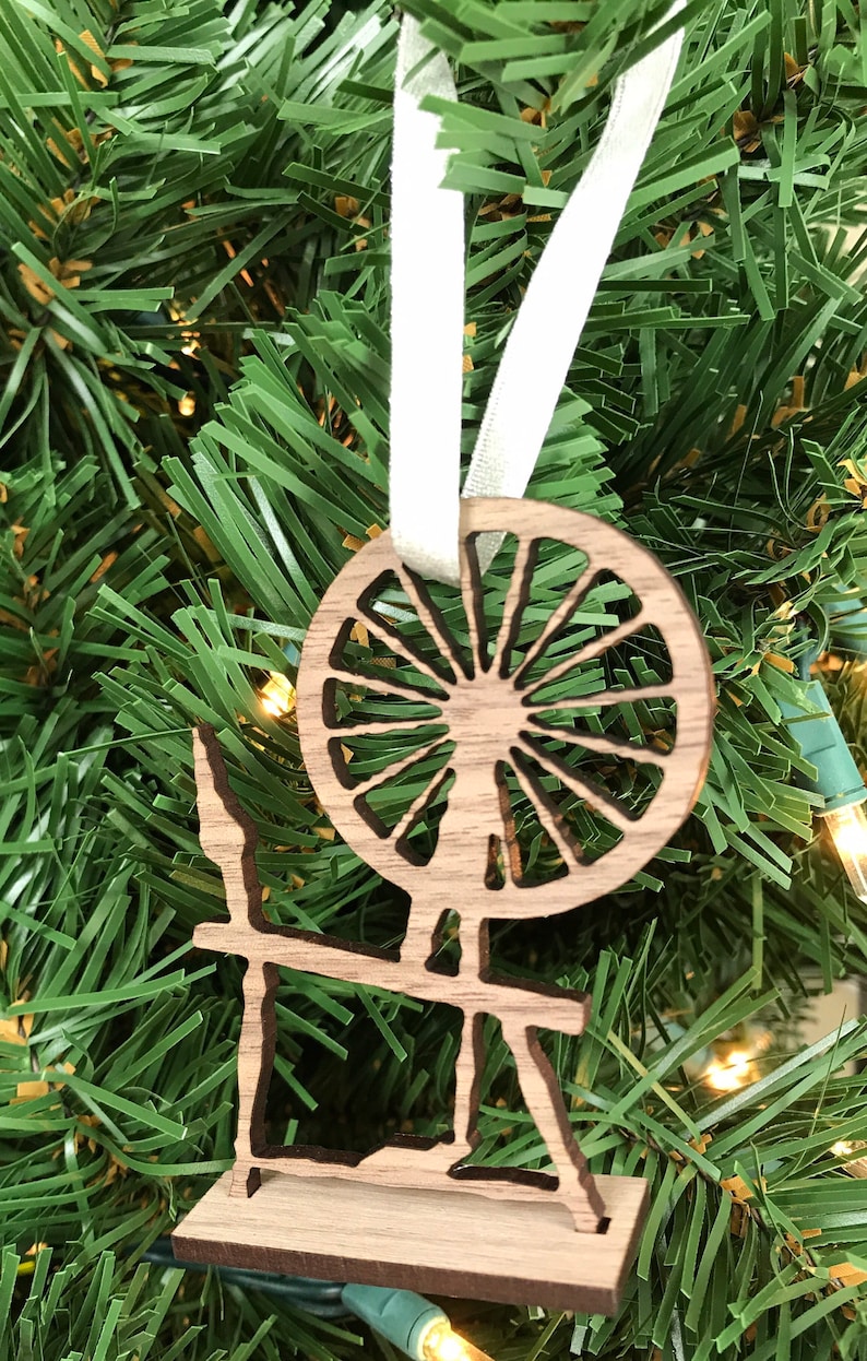 May include: A wooden spinning wheel ornament with a white ribbon for hanging. The wheel is detailed with spokes and a center hub. The ornament is a light brown color.