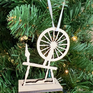 May include: A wooden spinning wheel ornament with a white ribbon hanging from the top. The ornament is a silhouette of a spinning wheel with a large wheel and a base.