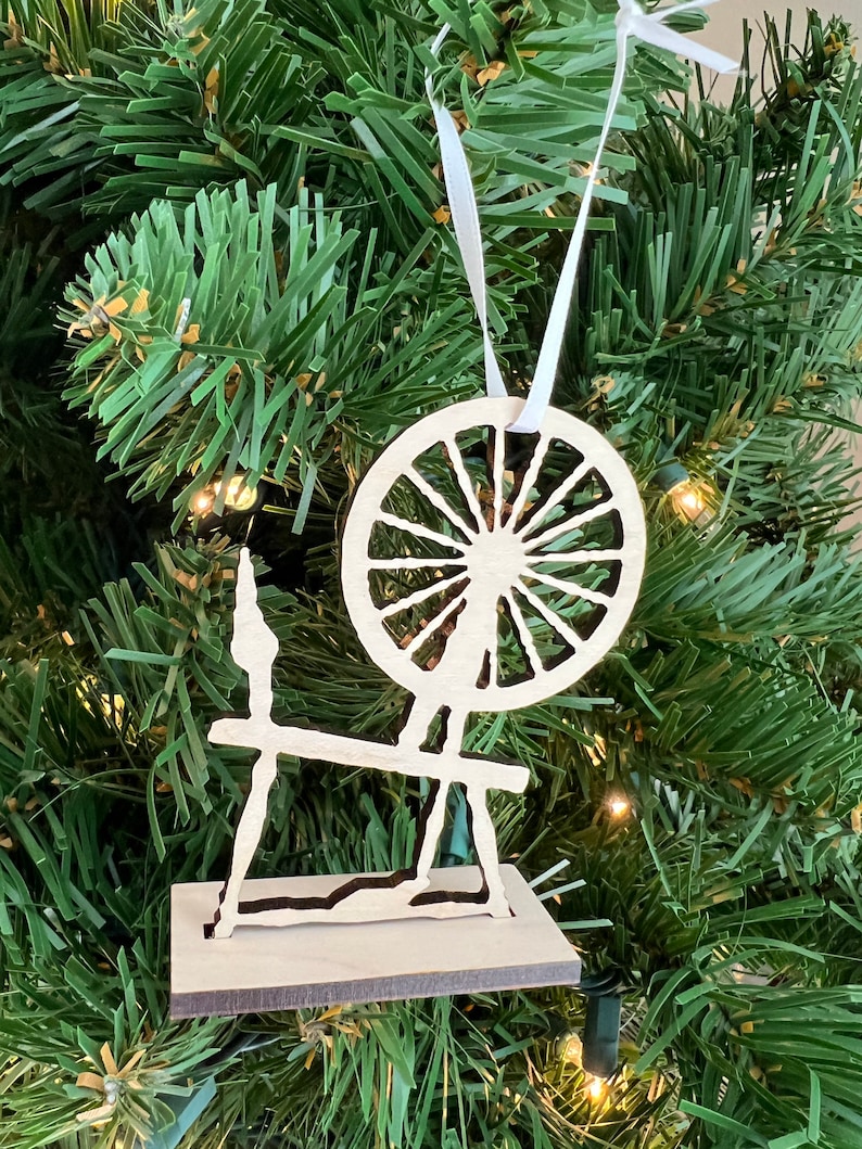 May include: A wooden spinning wheel ornament with a white ribbon hanging from the top. The ornament is a silhouette of a spinning wheel with a large wheel and a base.