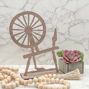 May include: Wooden spinning wheel silhouette with a brown finish. The wheel is set on a white marble surface with a small potted succulent plant and a wooden bead garland.