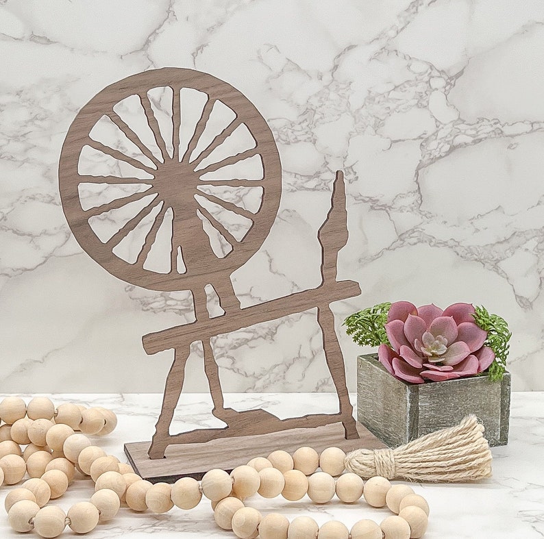 May include: Wooden spinning wheel silhouette with a brown finish. The wheel is set on a white marble surface with a small potted succulent plant and a wooden bead garland.