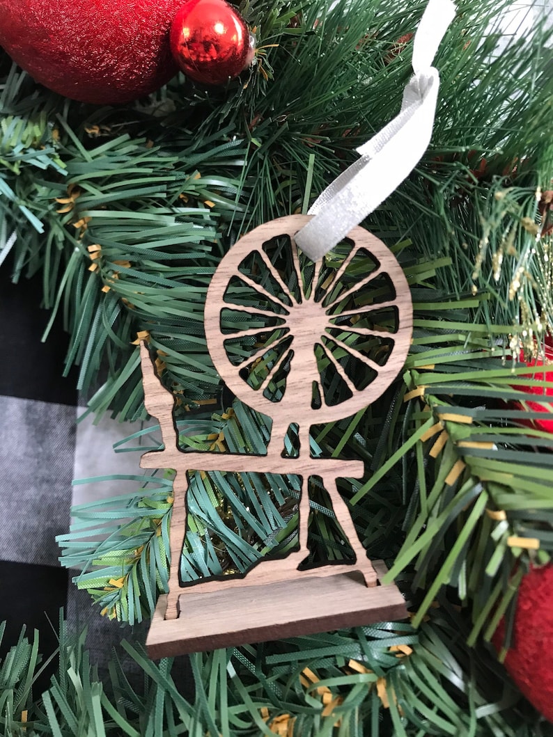 May include: Wooden Christmas ornament shaped like a spinning wheel. The ornament is brown and has a white ribbon attached to it.