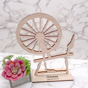 May include: Wooden spinning wheel cutout with a base that says "Arianni July 22, 2023". The spinning wheel is on a white marble surface next to a small potted succulent.