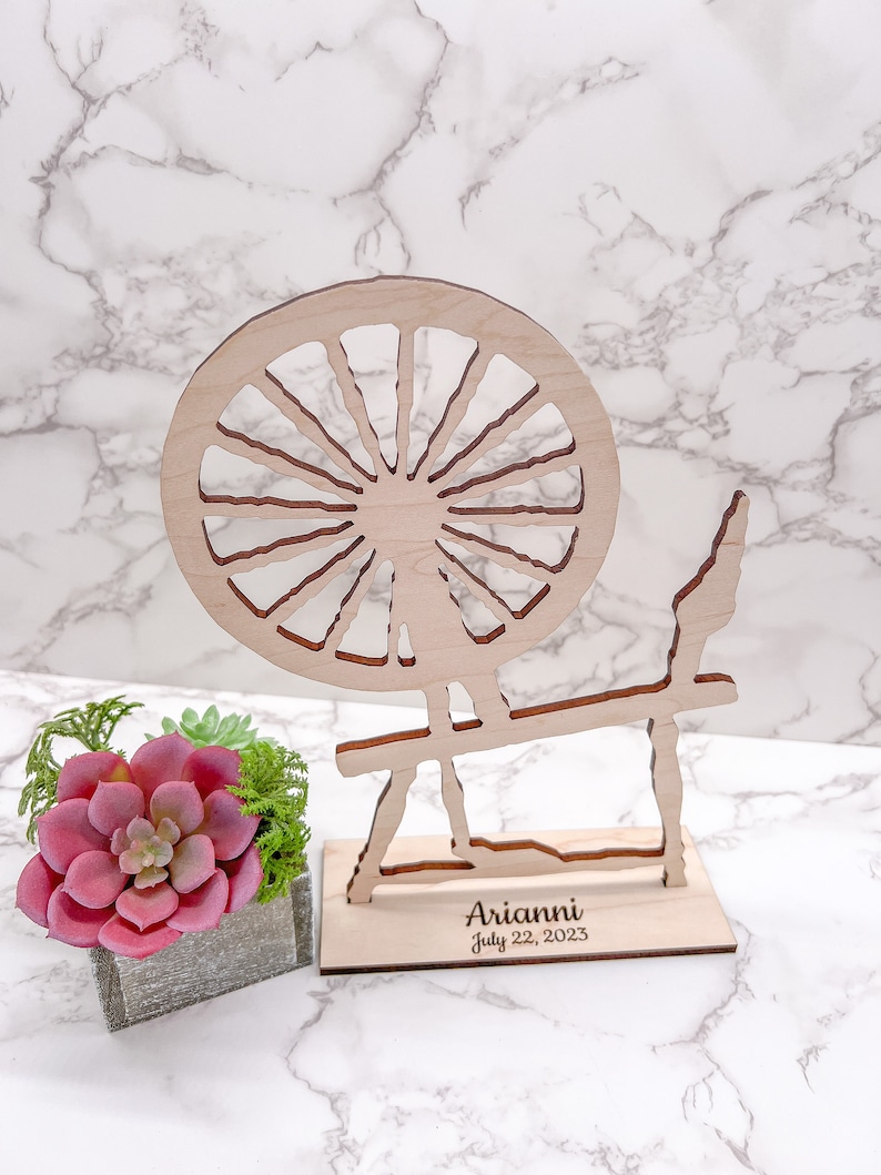 May include: Wooden spinning wheel cutout with a base that says "Arianni July 22, 2023". The spinning wheel is on a white marble surface next to a small potted succulent.