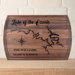 May include: A rectangular wooden cutting board with rounded edges. The board features the text "Lake of the Ozarks" and a map outline of the lake. Below the map is the text "THE WILLIAMS" and coordinates. A compass rose is also included.
