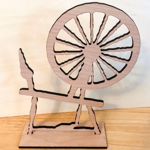 May include: A wooden spinning wheel with a large, round wheel and a stand. The wheel is made of light wood and has a dark brown outline. The stand is also made of light wood and has a dark brown outline.