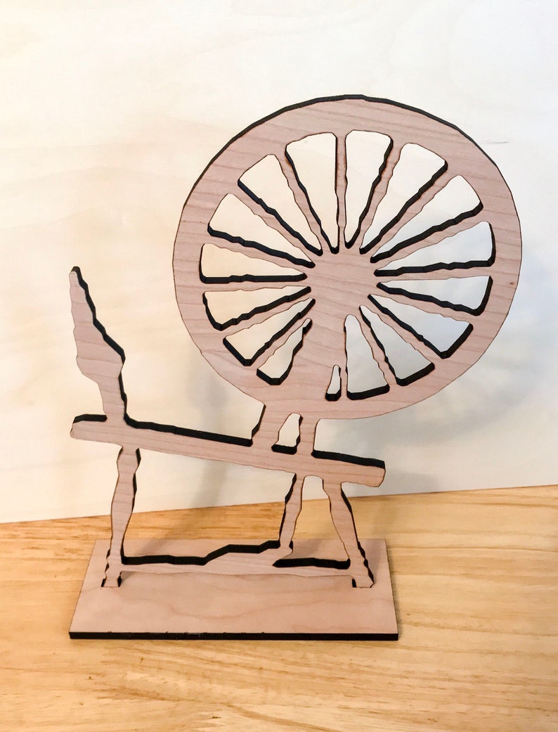 May include: A wooden spinning wheel with a large, round wheel and a stand. The wheel is made of light wood and has a dark brown outline. The stand is also made of light wood and has a dark brown outline.