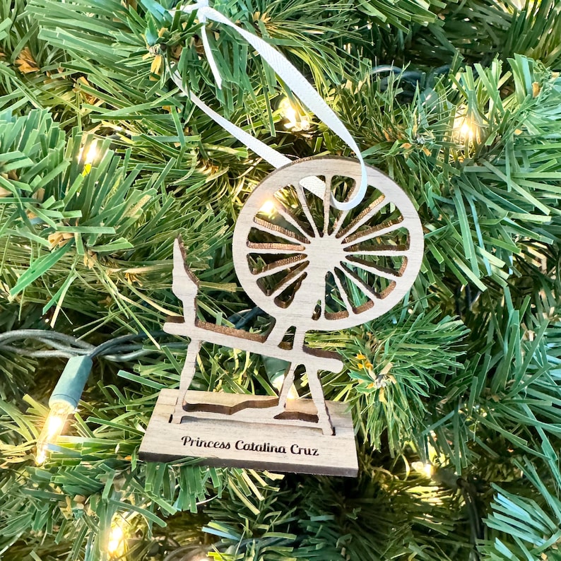 May include: A wooden spinning wheel ornament with a white ribbon hanging from the top. The ornament is decorated with the text "Princess Catalina Cruz".
