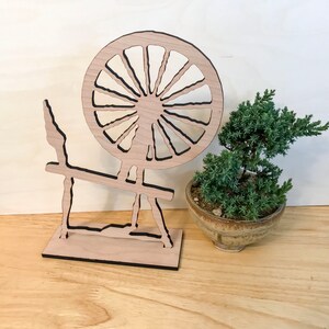 May include: A wooden spinning wheel with a black outline, standing on a wooden base. A small green potted plant is in the foreground.