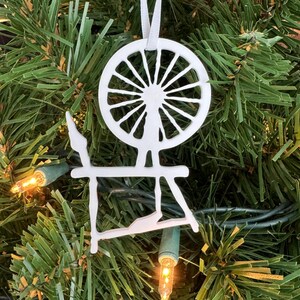 May include: White wooden spinning wheel ornament hanging on a green Christmas tree. The ornament is a silhouette of a spinning wheel with a large wheel and a stand.
