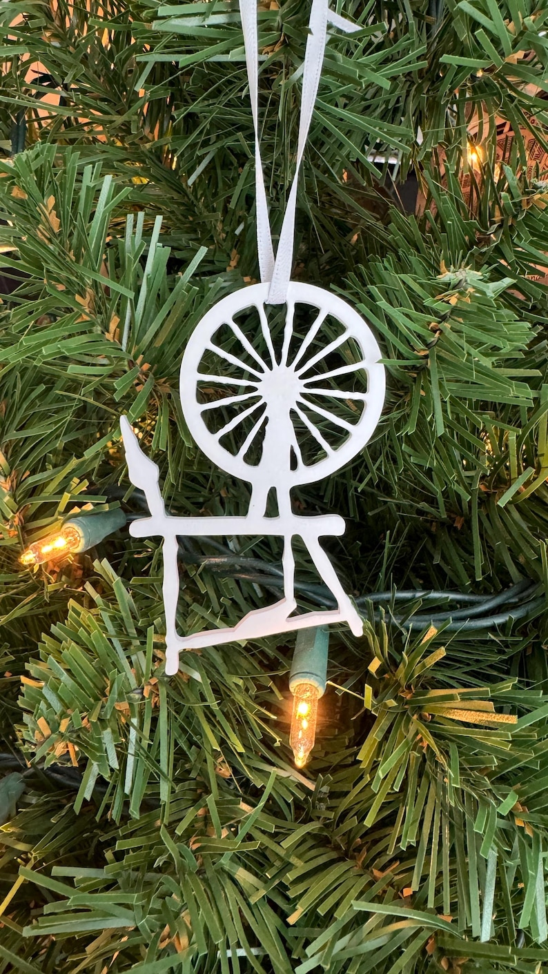 May include: White wooden spinning wheel ornament hanging on a green Christmas tree. The ornament is a silhouette of a spinning wheel with a large wheel and a stand.