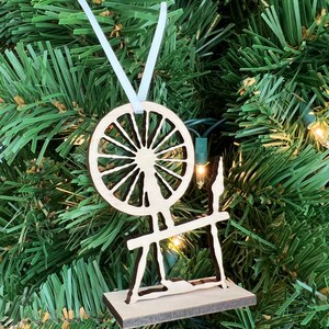 May include: A wooden spinning wheel ornament with a white ribbon for hanging. The wheel is cut out and the design is painted black.