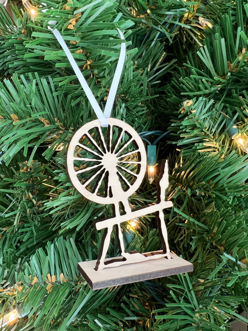 May include: A wooden spinning wheel ornament with a white ribbon for hanging. The wheel is cut out and the design is painted black.