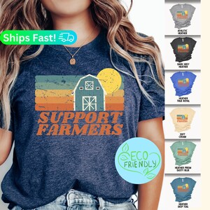 May include: A blue t-shirt with a vintage-style graphic of a barn and sunset. The text "Support Farmers" is printed below the graphic. The shirt is available in a variety of colors and is eco-friendly.