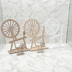 May include: Three wooden spinning wheels, two are brown and one is white. The wheels are all decorated with spokes and are mounted on stands. The stands are all different shapes and sizes.