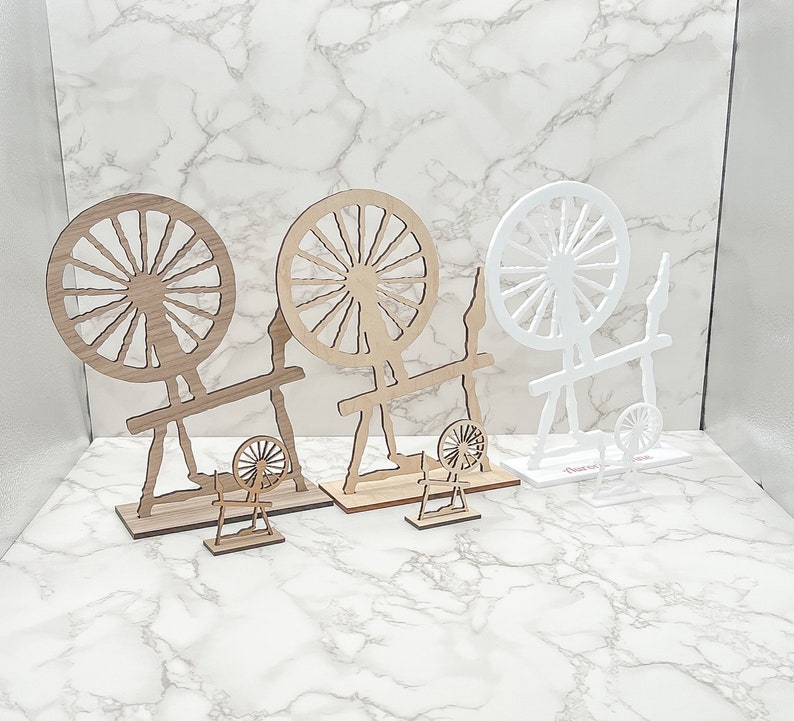 May include: Three wooden spinning wheels, two are brown and one is white. The wheels are all decorated with spokes and are mounted on stands. The stands are all different shapes and sizes.