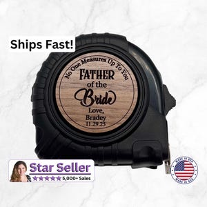 May include: A black retractable tape measure featuring a circular wood-grain inlay on the side. The inlay is engraved with the text: No One Measures Up To You, FATHER of the Bride, Love, Bradey, 11.29.25. The device has a textured black casing.