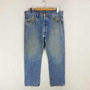 May include: A pair of light blue denim jeans with a faded wash, featuring a classic five-pocket design, a button fly closure, metal rivets, and belt loops, hanging from a wooden hanger against a plain, neutral-colored background.