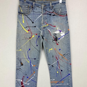 Puede incluir: Vaqueros de mezclilla azul claro con un diseño de salpicaduras de pintura multicolor que incluye trazos amarillos, rojos, negros, blancos, azules y naranjas. Los pantalones tienen cinco bolsillos, cierre de botones y bajos deshilachados.