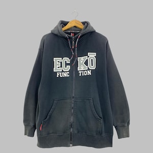 May include: A black zip-up hoodie featuring a hood with drawstrings and a front zipper closure. The chest displays the white and grey text ECKŌ FUNCTION in a bold, block-style font. It includes two front pockets and ribbed cuffs.