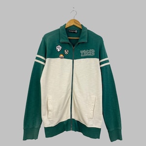 Puede incluir: Una chaqueta con cremallera en bloques de color verde y blanco roto, con mangas largas, cuello y dos bolsillos delanteros. El pecho presenta parches bordados y el texto T&CSD. Las mangas tienen dos rayas horizontales blancas cerca del hombro.