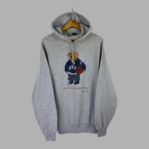 May include: A heather grey long-sleeve hooded sweatshirt featuring a graphic of a teddy bear wearing a blue USA hoodie and holding a basketball. The text Polo Bear by Ralph Lauren is printed below the bear, with a Polo Ralph Lauren label inside.