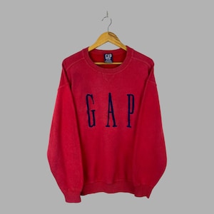 May include: A red long-sleeve crewneck sweatshirt featuring the word GAP embroidered in dark blue across the chest. The garment has a ribbed collar, cuffs, and hem, and includes a small GAP brand label on the inside of the neckline.