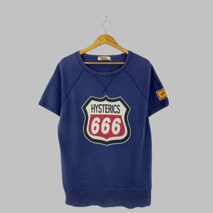 May include: A short-sleeve blue sweatshirt featuring a graphic on the chest shaped like a road sign with the text HYSTERICS and 666 in red and white. A small yellow rectangular patch with the text HE is visible on the right sleeve area.