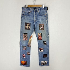 May include: Light blue denim jeans featuring a button fly and multiple rectangular patches with various graphic prints, including the word Oasis, stitched onto the front of both legs. The jeans have a classic five-pocket design and a medium wash.