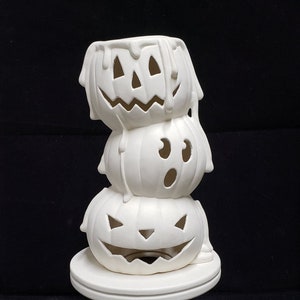 Könnte beinhalten: Ein weißer Kerzenhalter aus Keramik in Form von drei übereinander gestapelten Kürbissen mit unterschiedlichen Gesichtsausdrücken. Der untere Kürbis hat ein breites Lächeln, der mittlere Kürbis hat einen überraschten Ausdruck und der obere Kürbis hat ein tropfendes Gesicht. Der Kerzenhalter steht auf einem weißen Sockel.