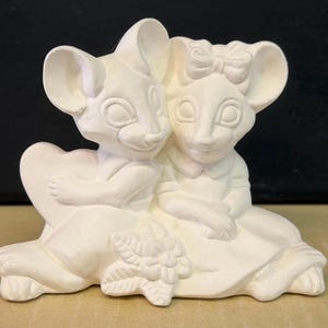 May include: A ceramic figurine of two mice embracing. One mouse has a bow on its head. They are sitting together on a heart-shaped base with a floral design. The figurine is unpainted and off-white.