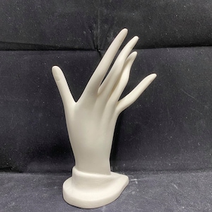 May include: White ceramic hand ring holder with a smooth, rounded surface. The hand is posed with the fingers extended and slightly curved, creating a space for a ring to be displayed.
