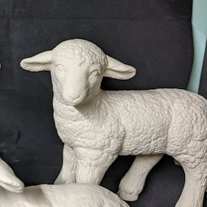Puede incluir: Figura de cordero de cerámica blanca con textura de lana detallada. El cordero está de pie, con la cabeza ligeramente girada hacia un lado. La figura no está pintada, mostrando el material cerámico en bruto. El fondo es oscuro.