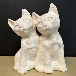 May include: Two white ceramic cat figurines sitting side by side. The cats are facing forward with their heads tilted slightly to the side. The cats have detailed features, including fur, eyes, and noses.