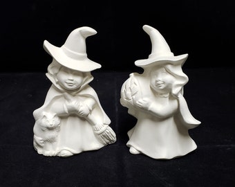 May include: Two white ceramic figurines of witches. One witch is holding a broom and a cat. The other witch is holding a pumpkin.