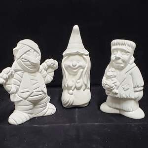 以下が含まれることがあります： 白いセラミック製のハロウィン人形3体。左の人形はミイラ、中央の人形は魔女、右の人形はフランケンシュタインの怪物です。