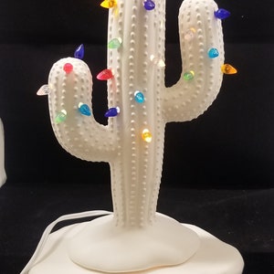 Könnte beinhalten: Weißer Keramik-Kaktus mit Beleuchtung. Der Kaktus hat einen Sockel und ist mit bunten Lichtern verziert.