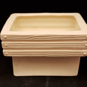 May include: A white ceramic planter with a square shape and a raised rim. The rim has a wood-like texture with three horizontal lines and small round accents.
