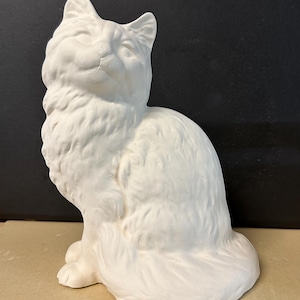 Pode incluir: Uma estatueta de gato de cerâmica branca em posição sentada. O gato tem uma aparência fofa com uma textura de pelo detalhada. A estatueta não está pintada e está sobre uma superfície neutra.
