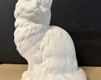 May include: A white ceramic cat figurine in a seated position. The cat has a fluffy appearance with detailed fur texture. The figurine is unpainted and sits on a neutral surface.