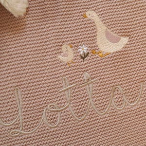 Puede incluir: Primer plano de una manta de punto rosa con detalles bordados. La manta presenta un ganso y un pollito, junto con una peque&ntilde;a flor. El nombre "Lotta" est&aacute; bordado en cursiva. La manta tiene un aspecto suave y texturizado.