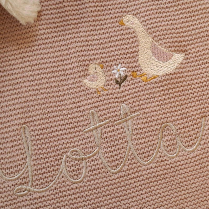 Puede incluir: Primer plano de una manta de punto rosa con detalles bordados. La manta presenta un ganso y un pollito, junto con una peque&ntilde;a flor. El nombre "Lotta" est&aacute; bordado en cursiva. La manta tiene un aspecto suave y texturizado.