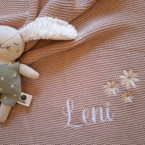 Könnte beinhalten: Eine gestrickte Babydecke in zartem Rosa, mit dem gestickten Namen "Leni" und floralen Akzenten. Ein Plüschhase mit weißem Ohr und grünem Outfit liegt auf der Decke. Die Decke hat eine strukturierte Optik.
