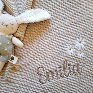 Könnte beinhalten: Eine beige gestrickte Babydecke mit dem Namen "Emilia" in brauner Schrift gestickt. Die Decke hat weiße Gänseblümchen-Stickereien und ein Stoffhasen-Spielzeug mit einem grünen Outfit mit Punkten.